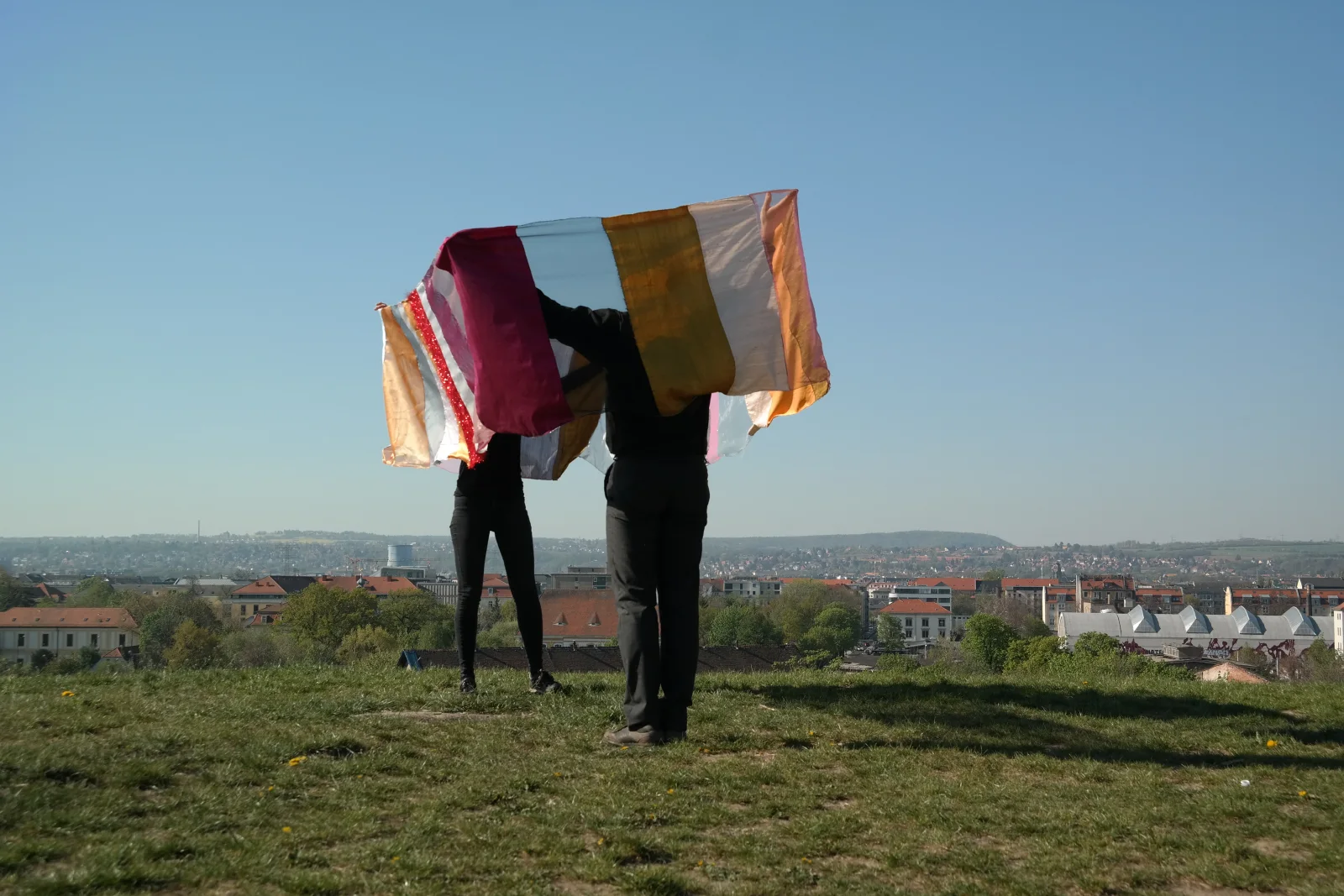 360° NOSW — zwei Personen halten ein langes, bunt gestreiftes Stoffband über ihre Köpfe, das ein Rechteck um ihre Körper spannt, auf dem Trümmerberg im Ostragehege über den Dächern Dresdens