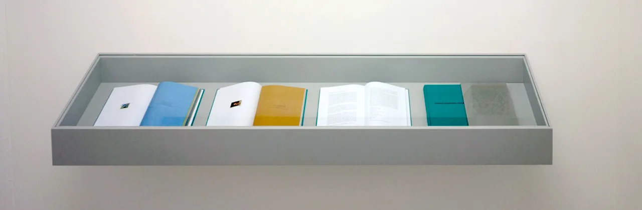 Mehrere Exemplare des Künstlerbuchs aufgeschlagen in einer Wandvitrine — türkise, blaue und goldgelbe Seiten nebeneinander