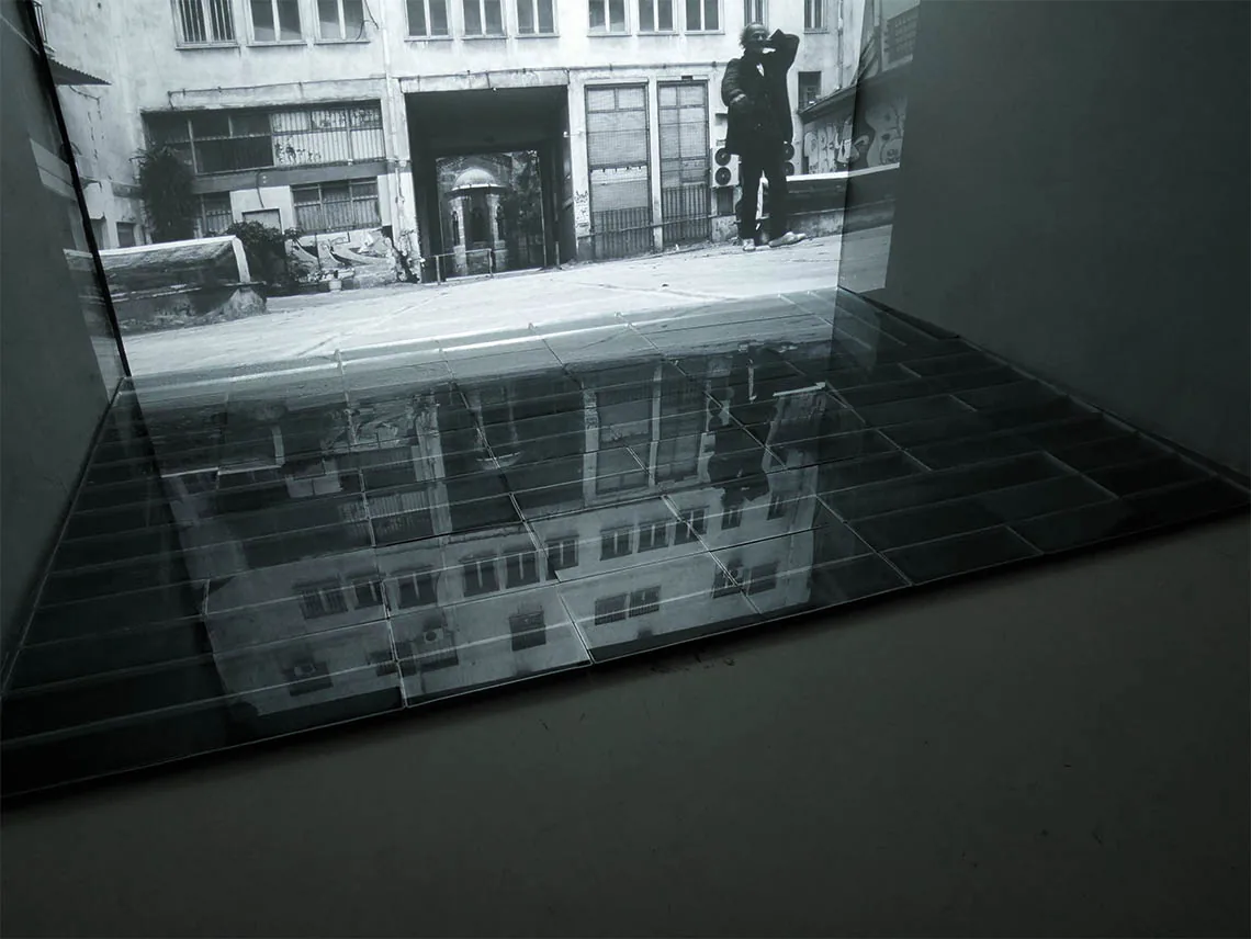 Condensate Mirror — die projizierte Athener Fassade kippt als Reflexion in die dunkle, kondensierte Spiegelfläche
