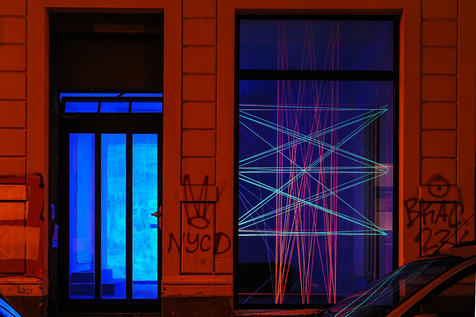 GROSSER PLAN (GG) — Schaufenster bei Nacht: im Schwarzlicht leuchtende rote und türkise Fäden überspannen das Fenster, daneben eine blau beleuchtete Tür