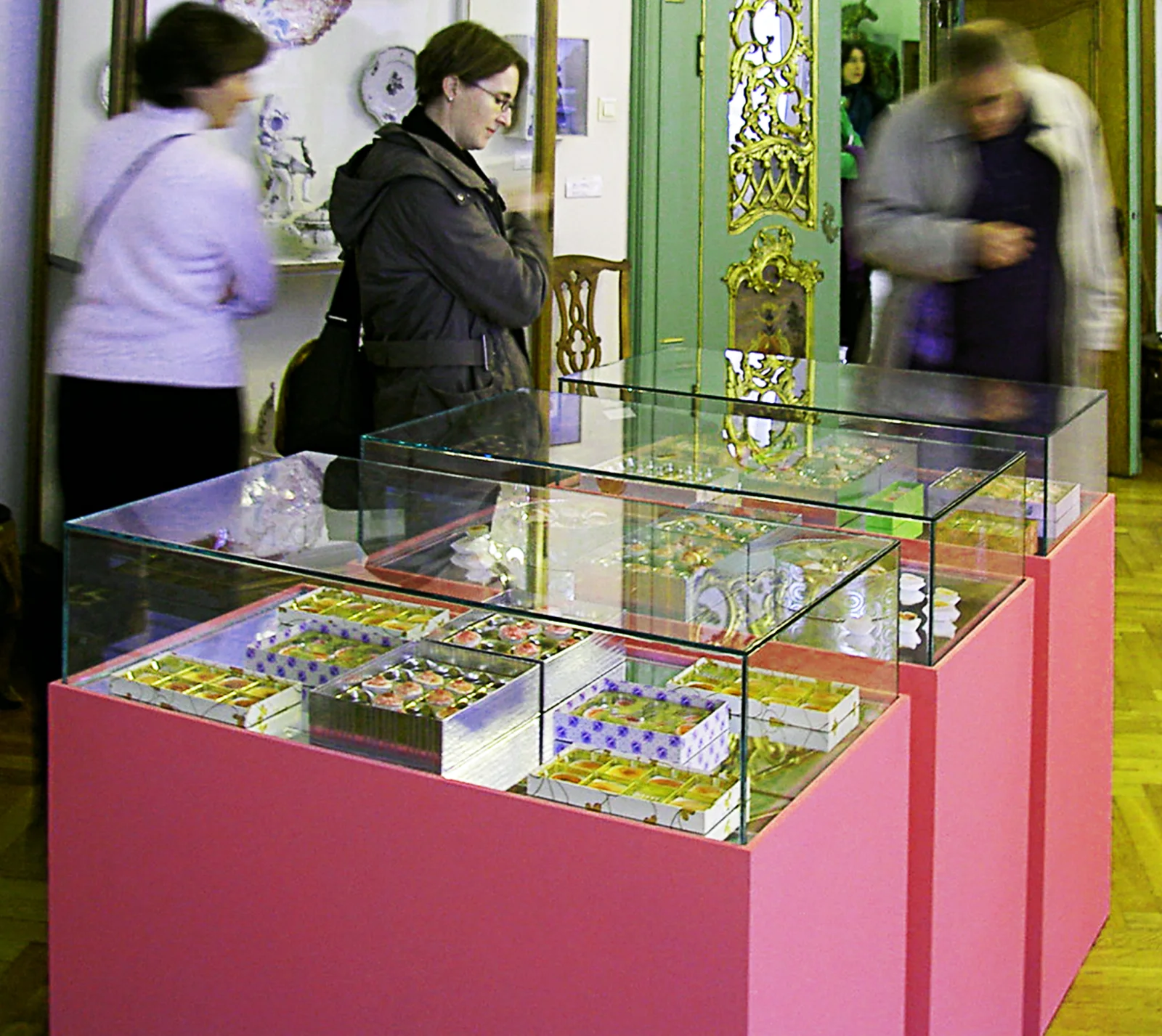 KANN LIEBE SÜNDE SEIN? — drei Spiegel-Vitrinen auf rosa Sockeln im Rokokosaal des Museumsberg Flensburg, mit Schachteln voller Pseudo-Pralinen, Besucher im Raum