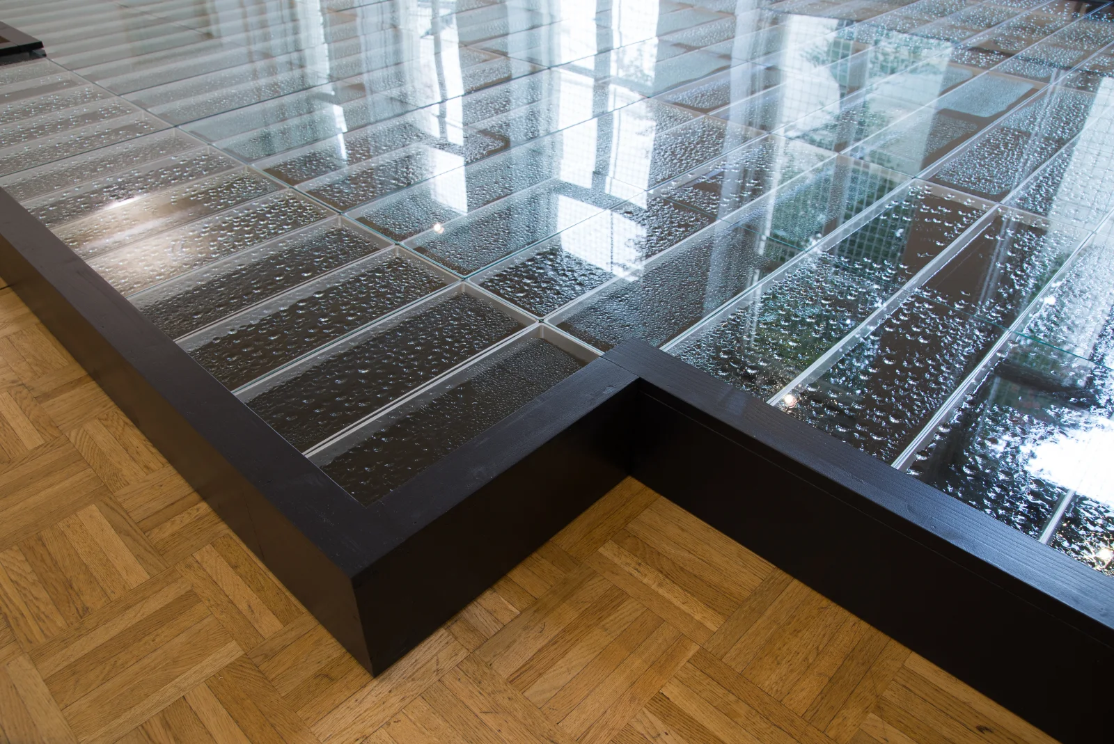Kondensat II — Detailansicht der Ecke der schwarz gerahmten Bodeninstallation auf Fischgrätparkett; unter den Glasplatten kondensiertes Tuschewasser mit unzähligen Tropfen, in den Scheiben spiegeln sich die Fenster des Saals. Kunstverein Bellevue-Saal, Wiesbaden 2014