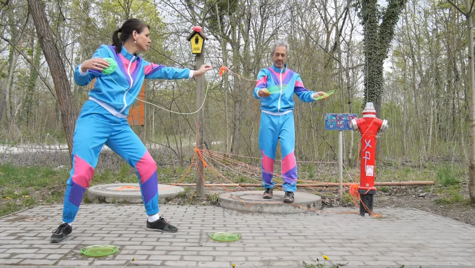 PLATEAUTANZ — zwei Personen in leuchtenden Trainingsanzügen auf je einem runden Podest, durch Schnüre verbunden; startet das Video