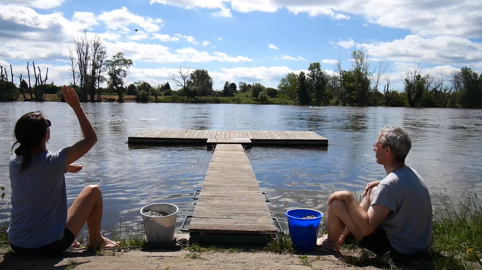 SCHOTTERVERSENKEN — Standbild: Birgit Schuh und Christoph Rodde sitzen am Elbufer zu beiden Seiten des Stegs, sie wirft Schotter auf den Bootsanleger im Fluss; startet das Video