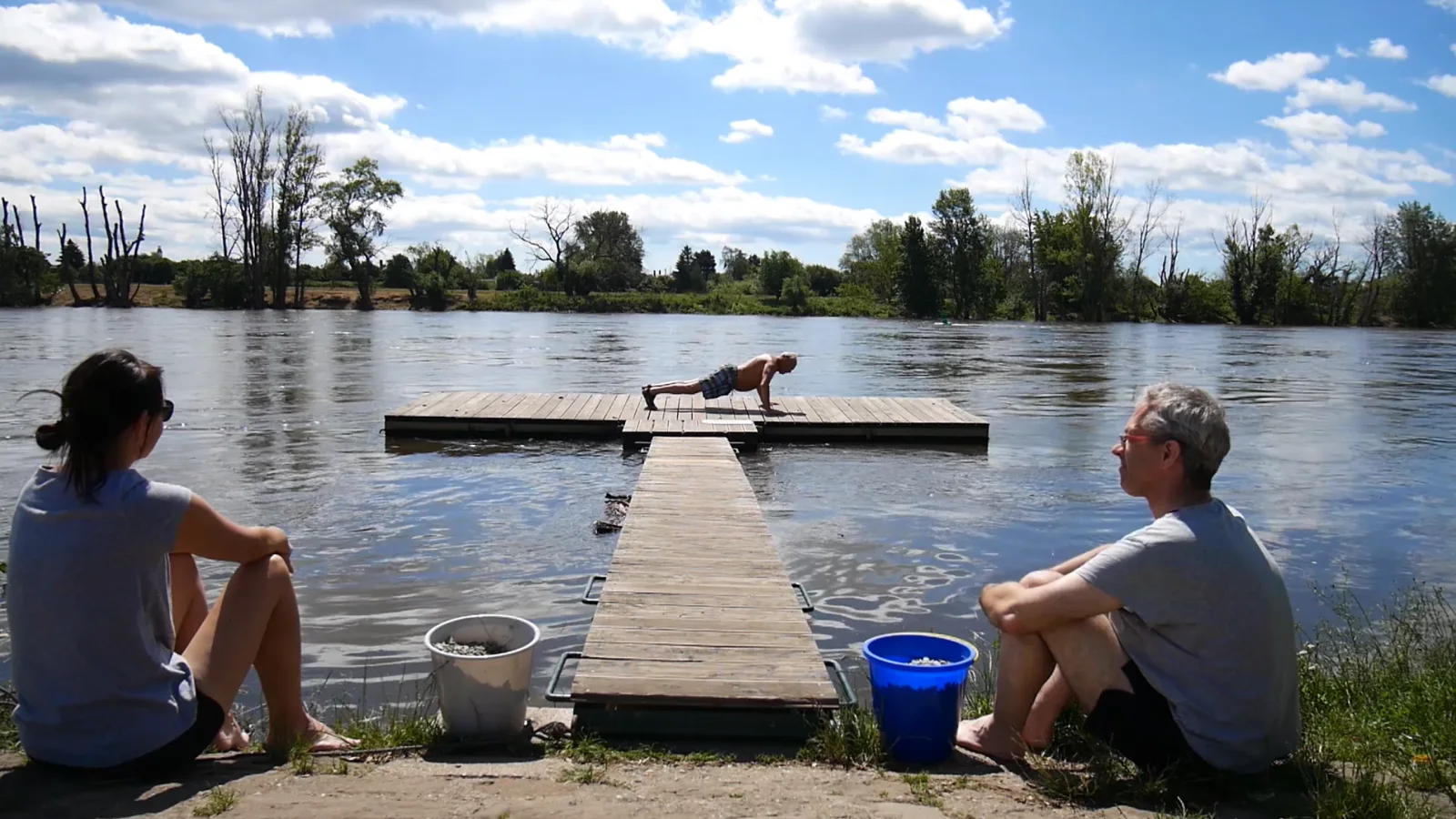 SCHOTTERVERSENKEN — ein Fremder macht Liegestütze auf dem Bootsanleger, die beiden Performer sitzen am Ufer, Standbild aus dem Video