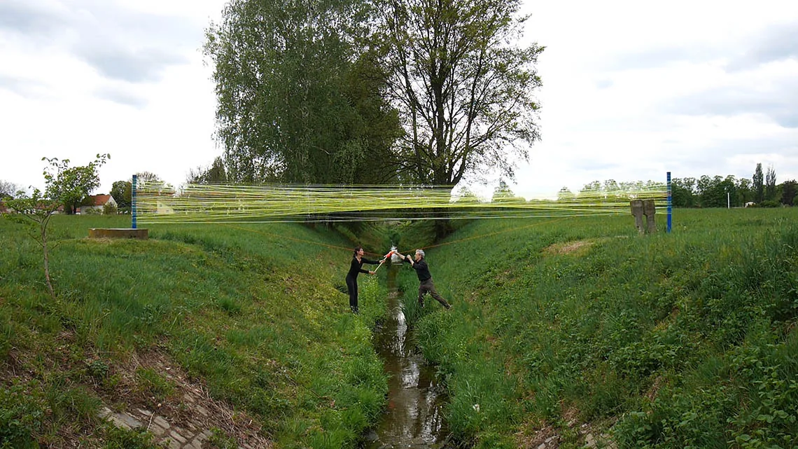 zusammen rumspinnen — Performance von Schuh/Rodde über einen Bachlauf: zwischen zwei blauen Pfosten gesponnene Fäden, die beiden Spinnenden vom Wasserlauf getrennt.
