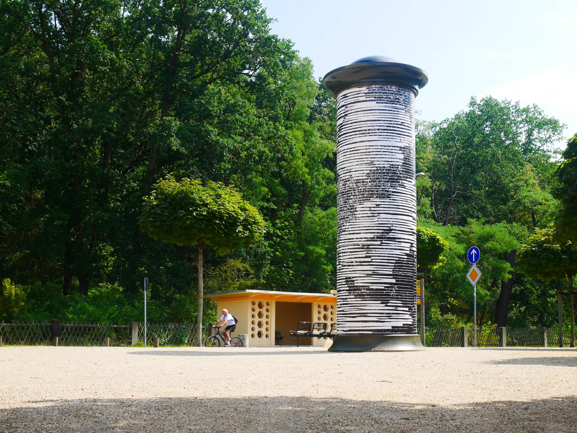 CHIFFRE#CLOUD — die plakatierte Litfaßsäule steht auf einem Kiesplatz am Waldrand, ein Radfahrer passiert im Hintergrund. Kunst im öffentlichen Raum, Dresden, 2019