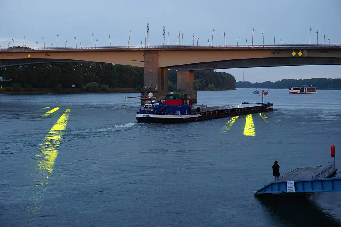 EINDUTZEND — gelbe Lichtspuren auf dem Rhein in der Dämmerung, ein Frachtschiff passiert die Brücke, Rheinufer Worms, 2022; öffnet das Video auf Vimeo