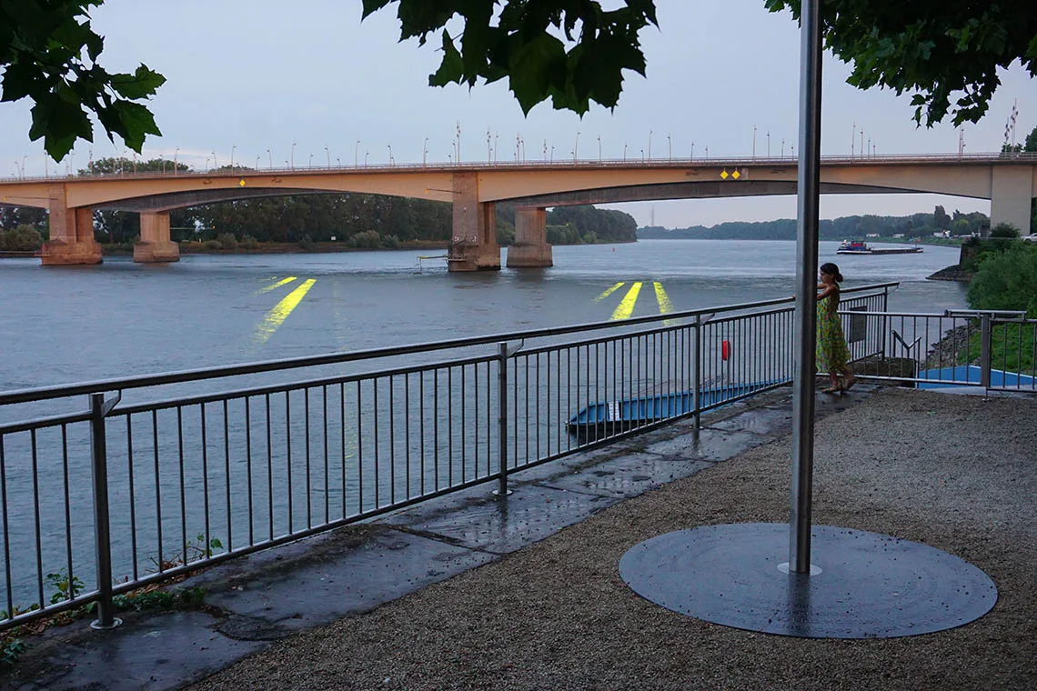 EINDUTZEND — der Aussichtspunkt am Rheinufer mit dem Mast der Hornlautsprecher; durch das Laub der Blick auf die goldgelben Lichtspuren im Wasser, Worms