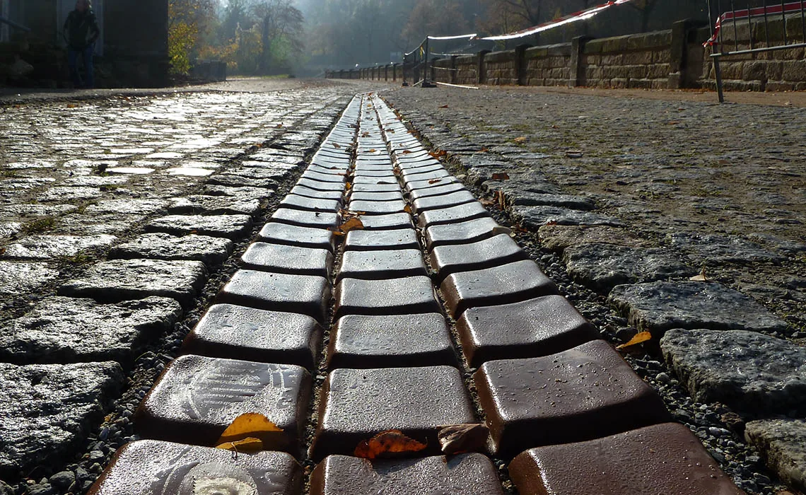 SCHOKOFLUSS — glänzende Rinne aus schokoladenfarbenem MMA-Beton im nassen Kopfsteinpflaster, Dresden, 2011