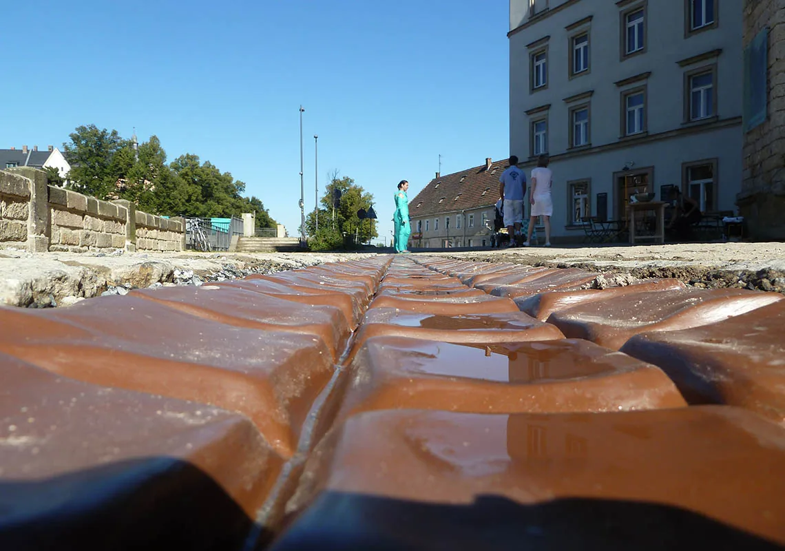 SCHOKOFLUSS — Detail der glänzenden Betonrinne mit Pfütze, im Hintergrund eine Passantin